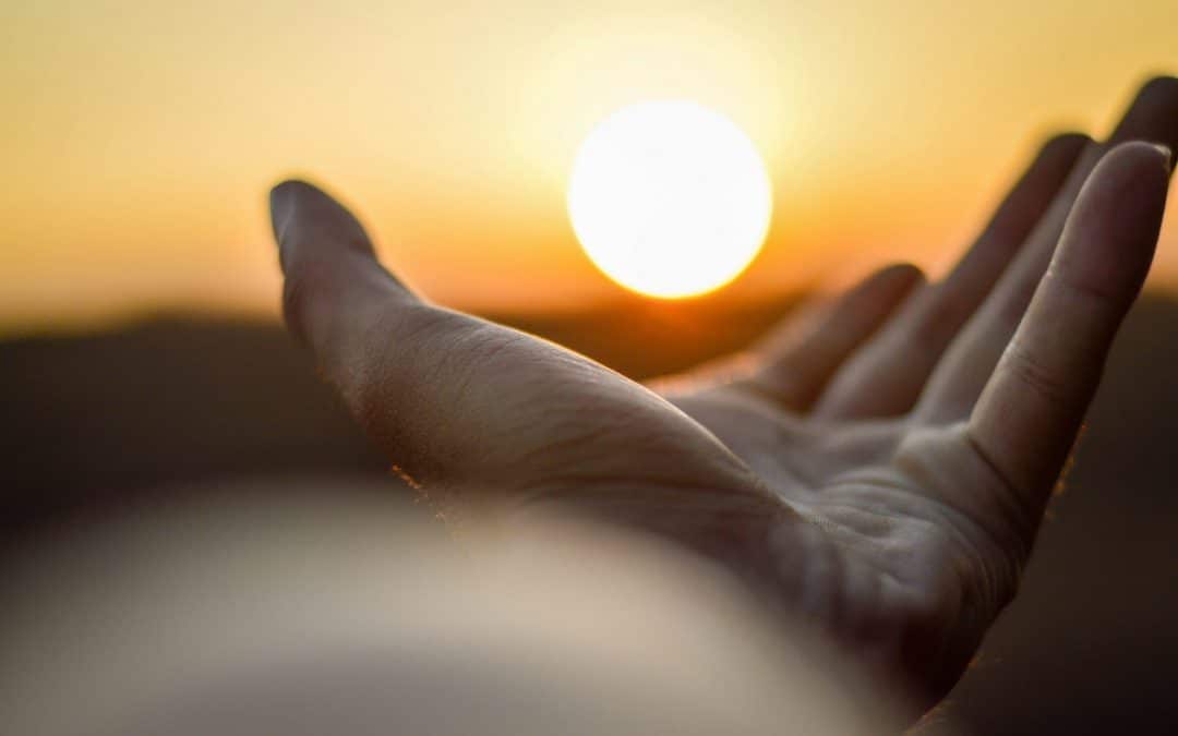Photograph showing an outstretched hand in the foreground and the setting sun in the background.